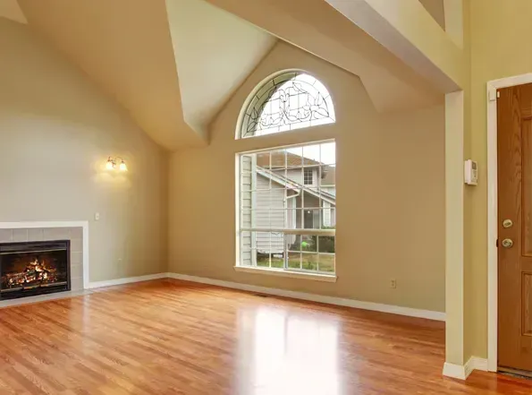 A bright living room with a fireplace, hardwood floors, and a large arched window overlooking a house.