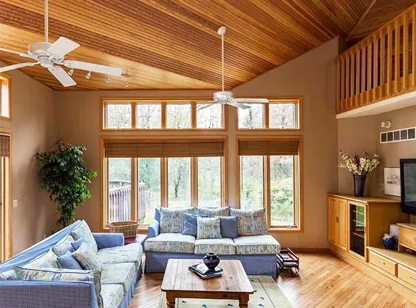 Living room with wood ceiling, beige walls, blue sofas, and large windows overlooking greenery. Two ceiling fans and a loft are also visible.