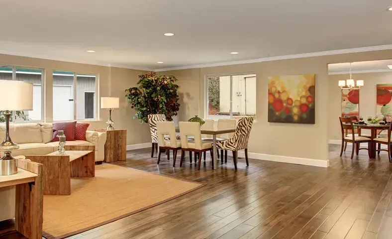 Spacious living room with hardwood floors, seating area, and dining table. Neutral walls, large windows, and colorful artwork.