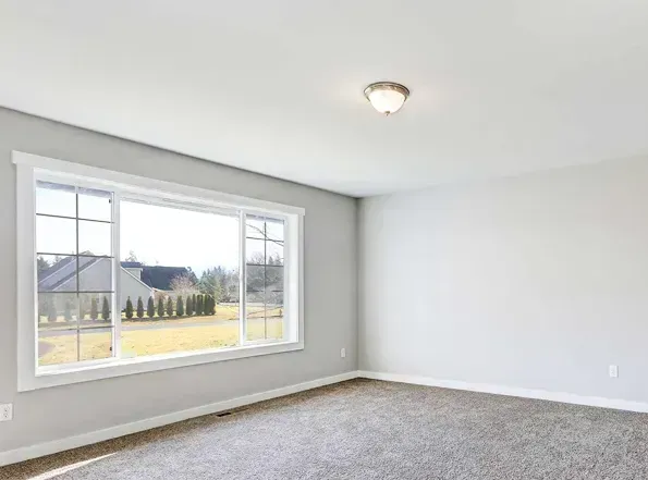 Empty room with large window overlooking a grassy yard and other homes. Gray carpet and light-colored walls.