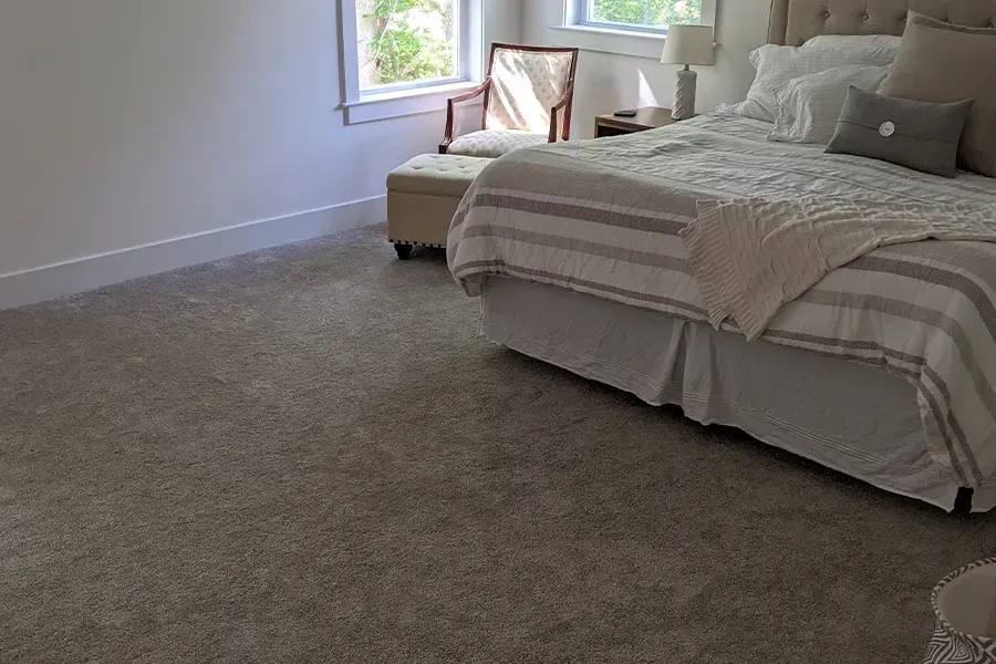 Bedroom with a large bed covered in white and gray bedding, small chair by the window, and light gray carpet.