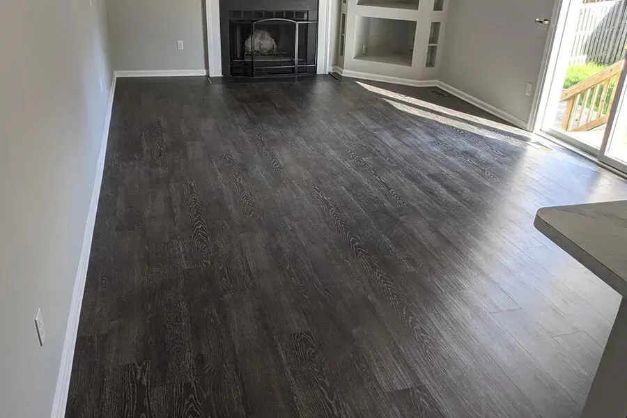 Dark wood-look flooring in a room with a fireplace and a door leading to a deck. Sunlight streams across the floor.