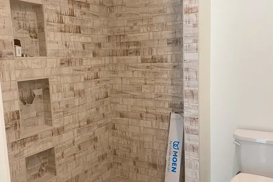 Bathroom with brick-style tile in the shower, featuring three built-in shelves. A roll of Moen shower trim rests nearby.