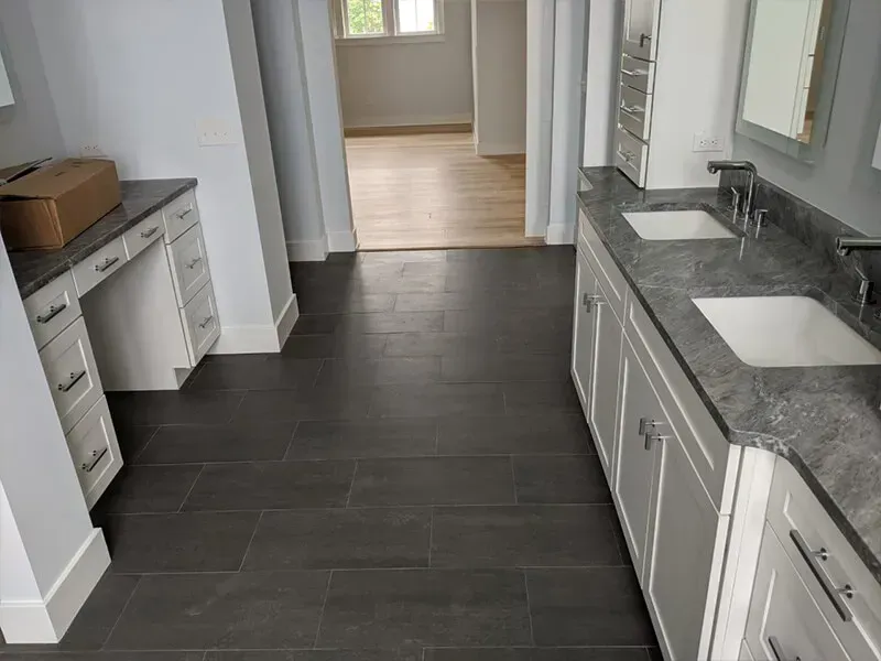 Bathroom with gray floor tiles, white vanity, and a gray countertop. Another room is visible through the doorway.