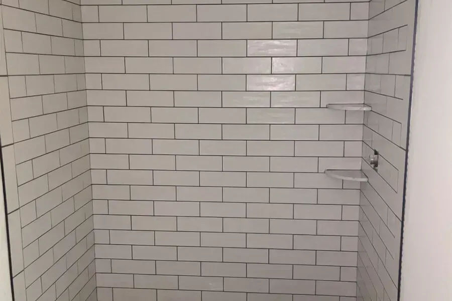 White tiled shower stall with dark grout, featuring two clear shelves.