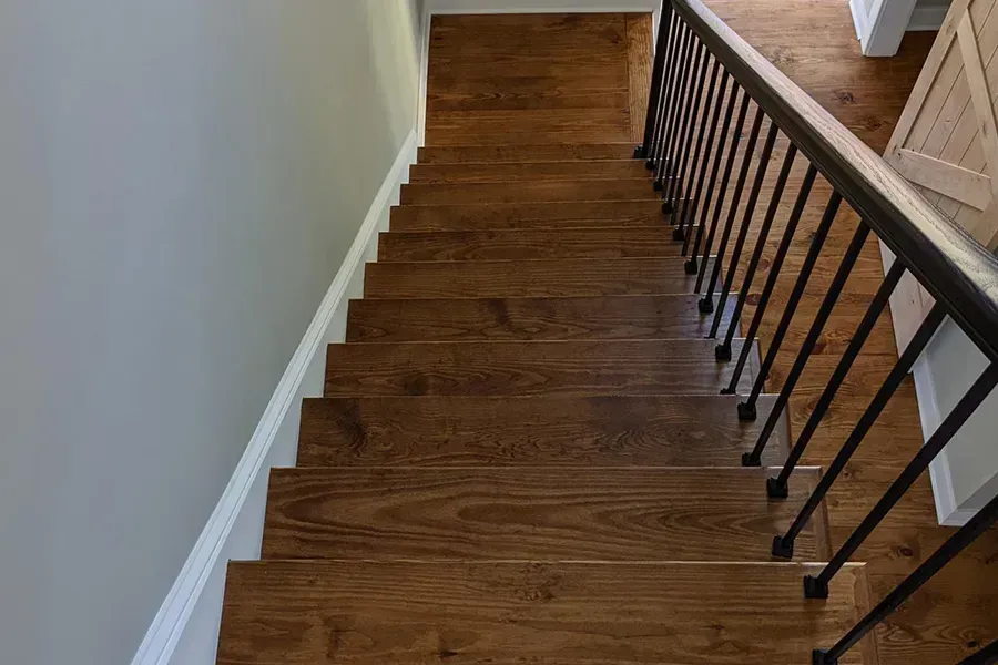 Wooden staircase viewed from above, with black metal railing and a white wall to the left.