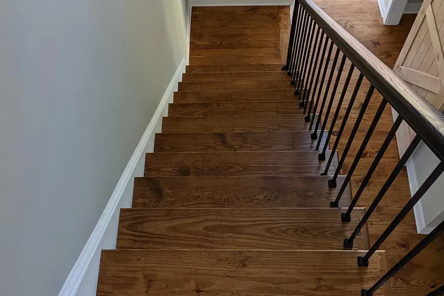 Wooden staircase with black railing descending from top right corner. Gray wall on the left.