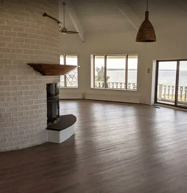 A spacious, empty living room with a whitewashed brick fireplace and large windows overlooking a body of water.