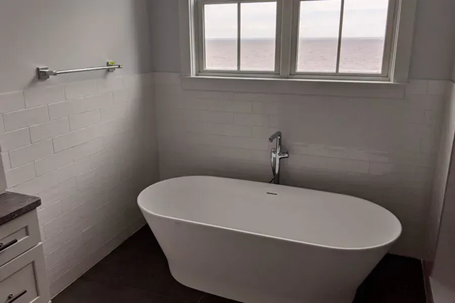 A modern bathroom with a white oval bathtub under a window. White subway tile walls and dark gray flooring.