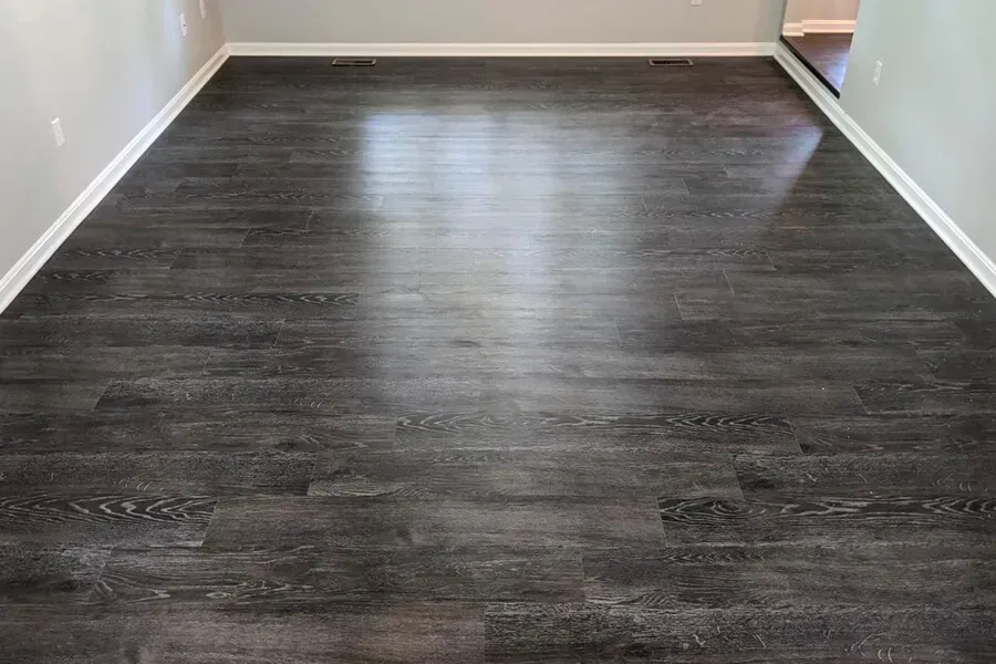 Dark wood-look flooring in a room, reflecting light. White trim lines the base of the walls.