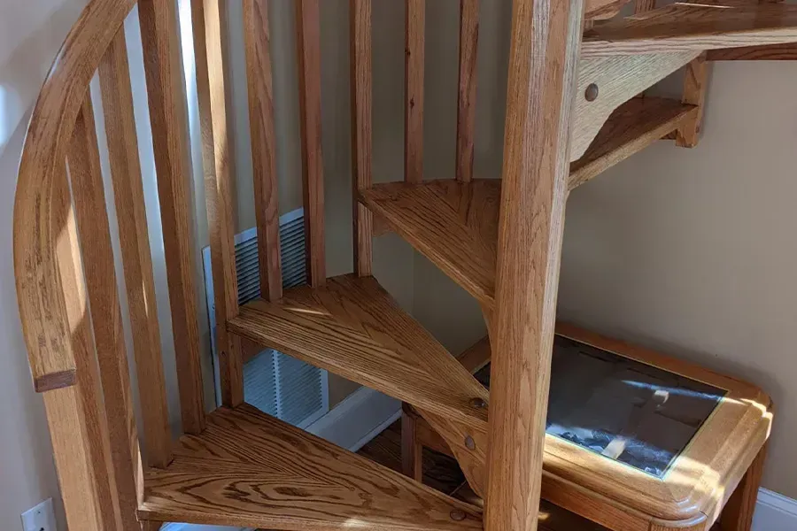 Wooden spiral staircase with a curved railing, next to a small shelf and wall.