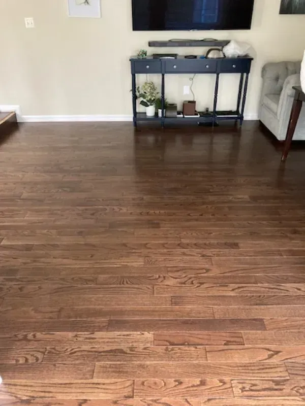 Hardwood floor with reddish-brown tones in a living room. A dark blue console table with plants and a TV above it are in the background.