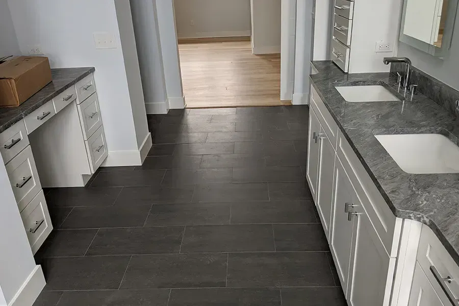 Bathroom with dark gray tiled floor, white cabinets, two sinks, and a vanity. A doorway leads to another room.
