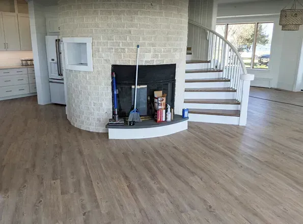 Newly remodeled interior with wood-look flooring, a brick-faced fireplace, and a curved staircase. A broom, vacuum, and cleaning supplies sit near the fireplace.