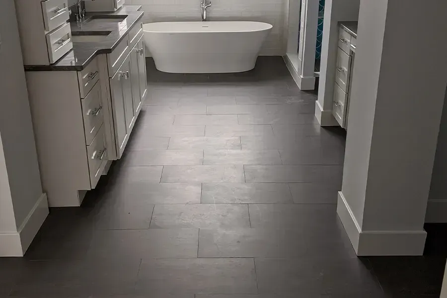 Dark wood-look tile floor in a modern bathroom. White cabinets line the walls. A white soaking tub is in the background.