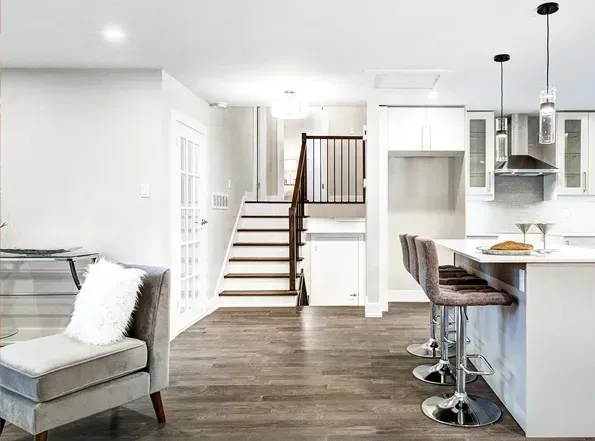 Modern kitchen and living area. Dark wood floors, white cabinets, and a staircase leading upstairs. Gray chaise lounge.