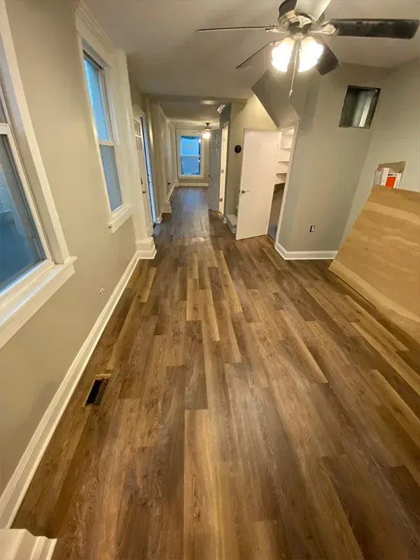 A hallway with wood-look flooring and tan walls. Windows and doors are along the hallway. A ceiling fan is visible.