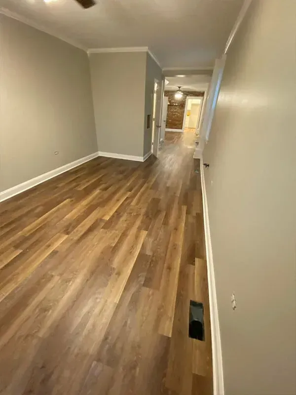 Empty room with wood-look flooring, gray walls, and white trim. A hallway leads to a brick wall in the distance.