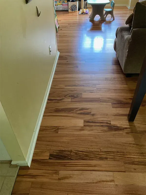 Wooden flooring extends from a beige wall into a room with furniture. The wood has various dark and light brown shades.