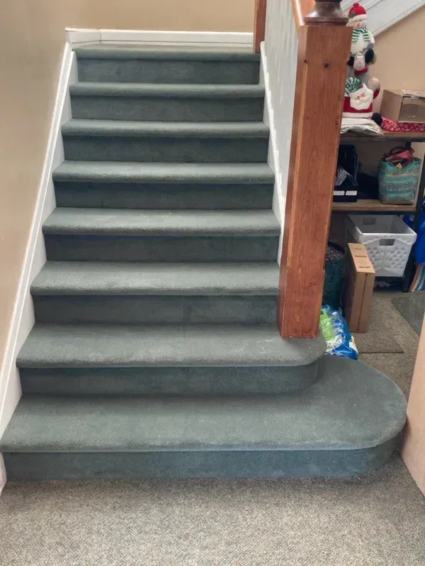 Staircase with green carpet, white trim, and wooden handrail leads up to a landing. The bottom stair has a rounded edge.