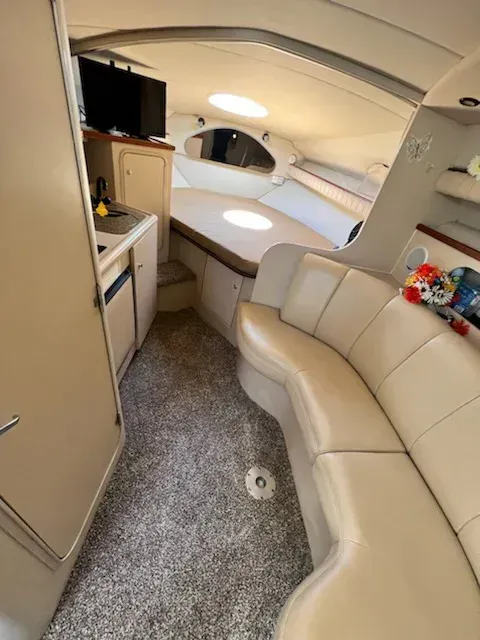 Interior of a boat cabin with a beige curved sofa, small kitchen area, and a bed at the front. Gray carpet covers the floor.