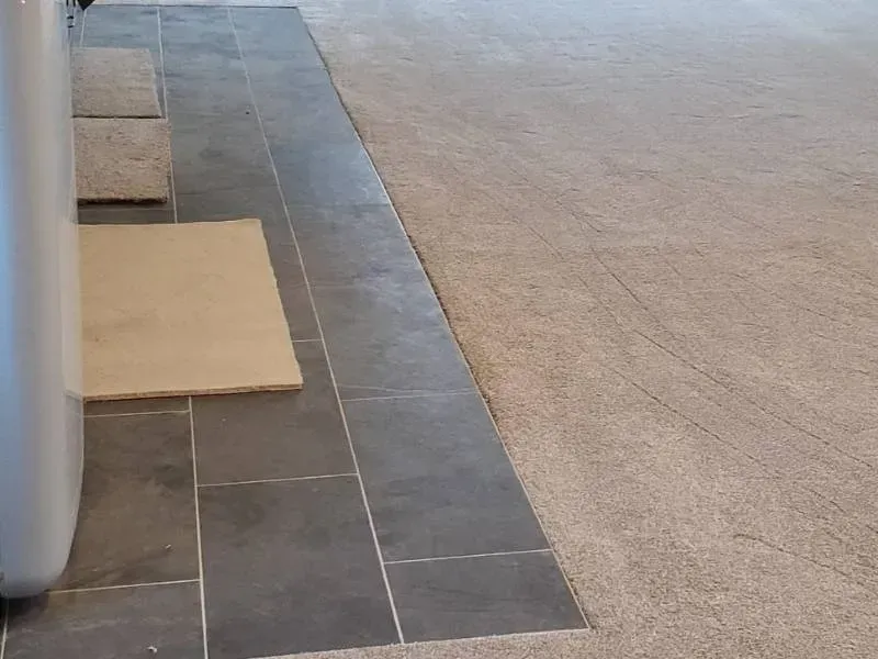 Dark gray tiled floor next to a light brown carpeted floor. A cardboard square sits on the tiles.
