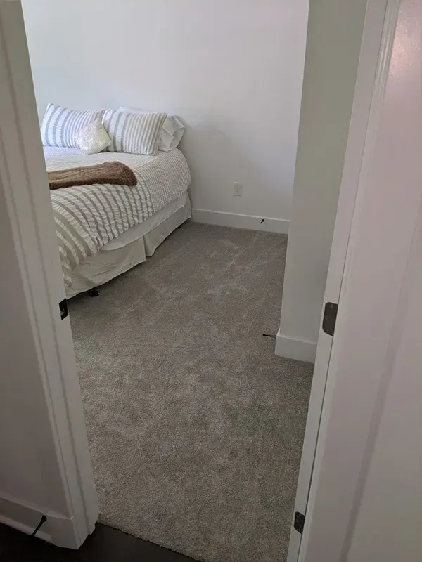 Bedroom interior with a bed covered in pillows and blankets; light gray carpet and white walls.