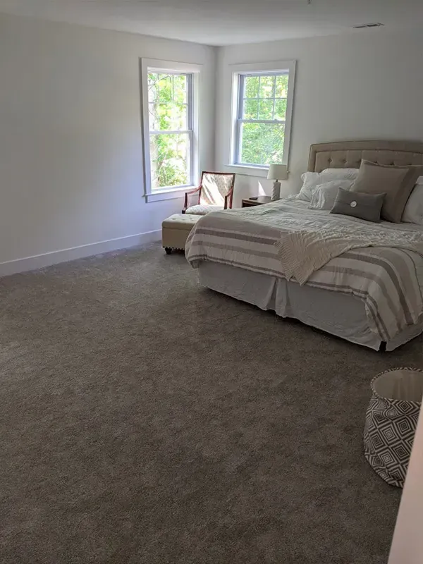 A bright bedroom with a bed, windows, and carpet. Sunlight streams in from the windows. A chair sits near the windows.