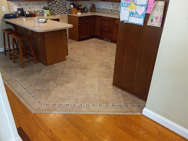 A kitchen with a wooden floor leading up to a tiled kitchen floor. There is a kitchen island and wooden cabinets.
