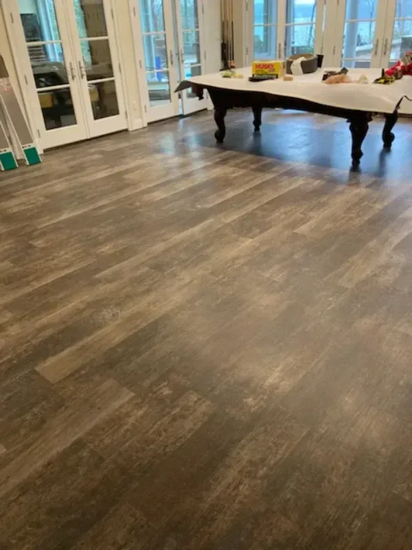 Brown and gray wood-look flooring in a room, leading toward a pool table. French doors in the background.