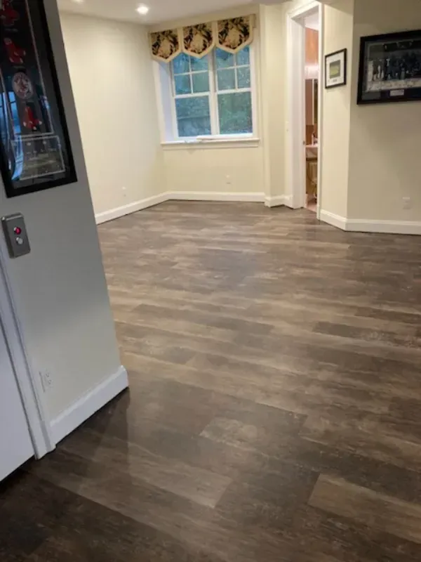 Empty room with dark wood-look flooring, a window with a valance, and light-colored walls. A doorway leads to a bathroom.