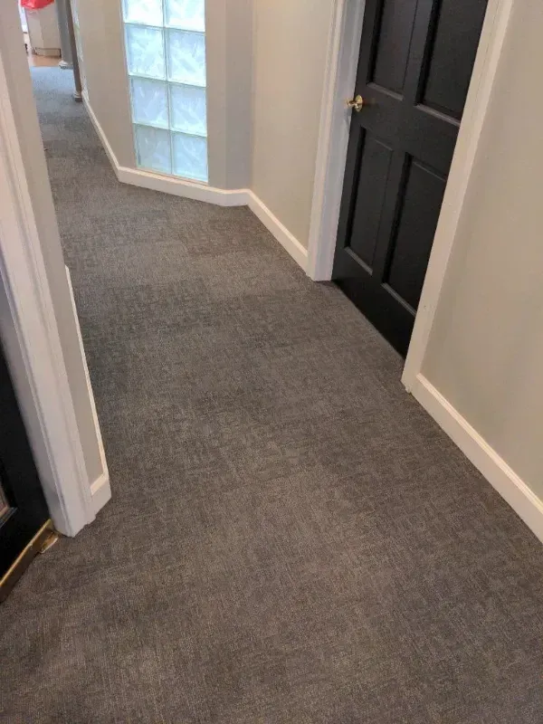 A hallway with gray carpet, white trim, gray walls, and a black door. A glass block window is visible.