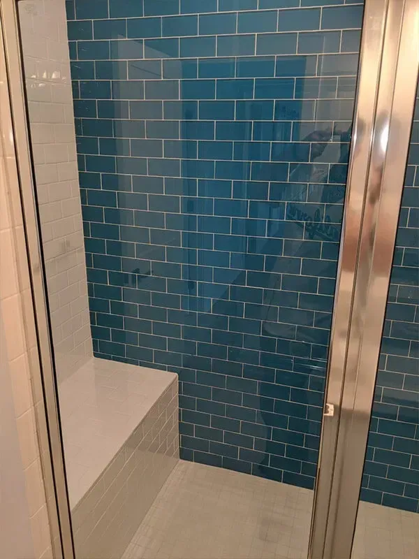 A shower interior with blue tile walls, a built-in bench, and a glass door.