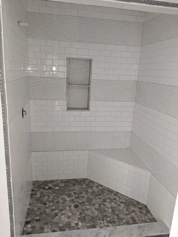 A tiled shower with a gray pebble floor, white subway tile walls, and a built-in bench. A niche is in the center.