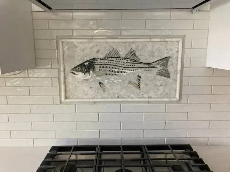 A white kitchen backsplash with a fish mosaic above a stovetop. The fish is black and gray, set within a white frame.