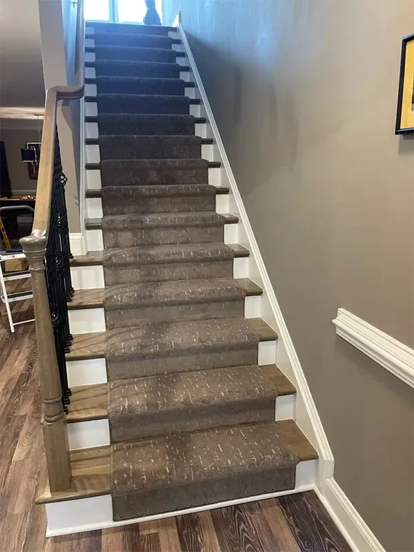 Staircase with brown carpet and white trim. Wooden banister with black iron details on the left, beige wall on the right.