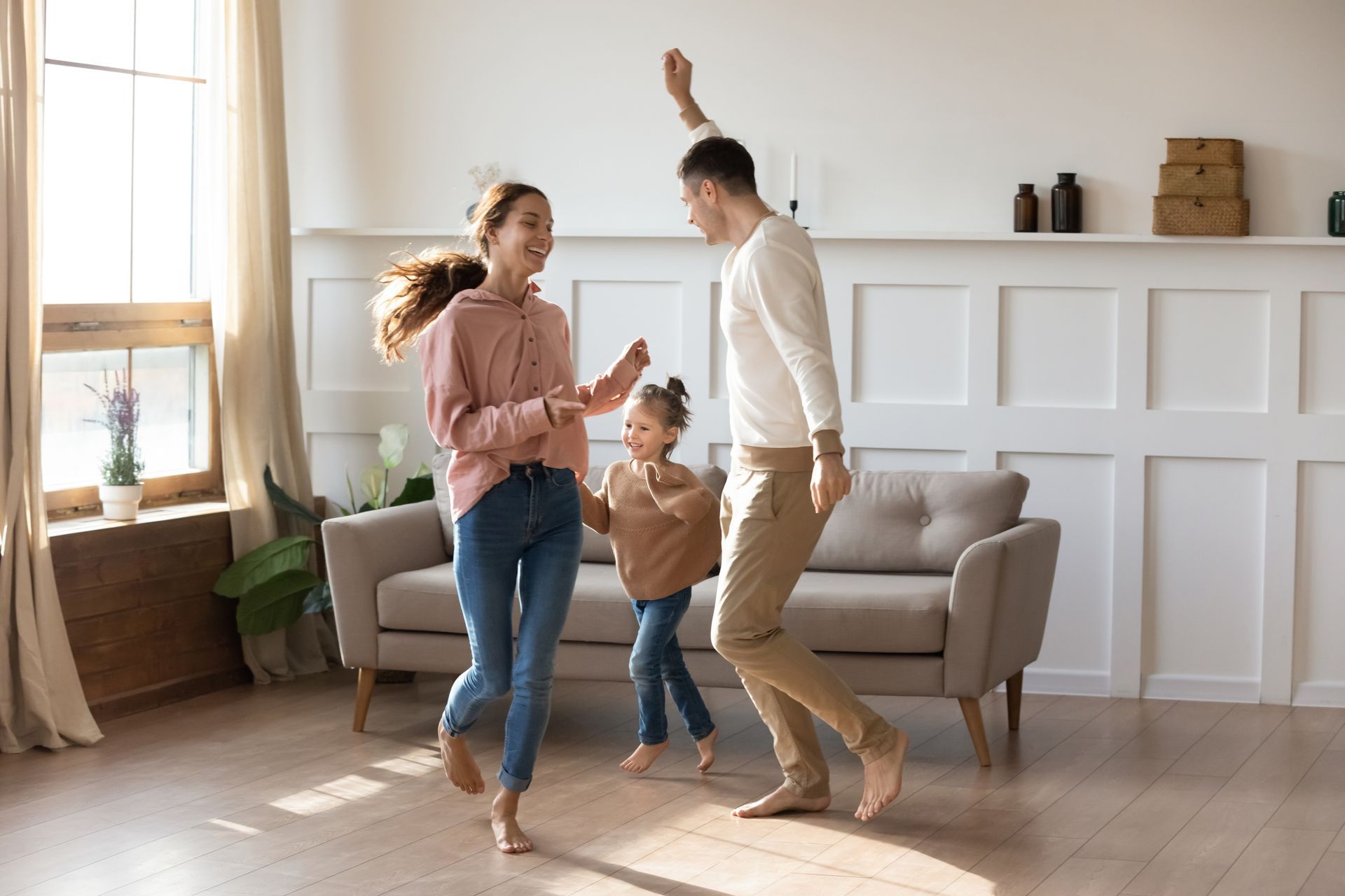 Family of three dancing joyfully in a living room with a large window. Smiling parents and a child have raised arms, celebrating.