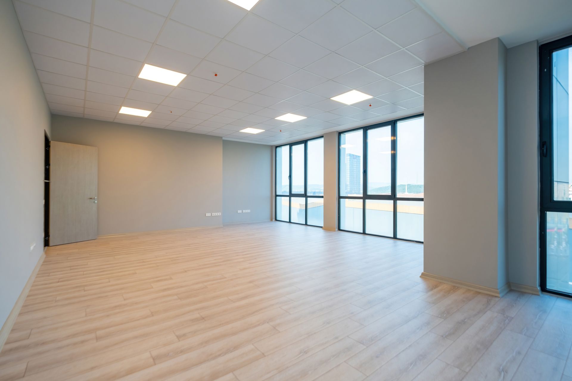 Empty office space with light wood flooring, large windows, and recessed ceiling lights. Walls are gray.