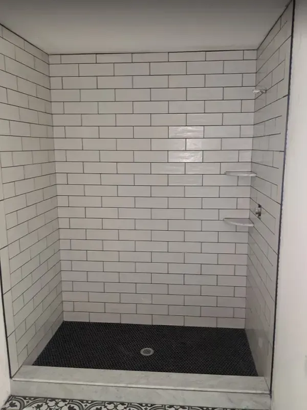 A white subway-tiled shower with a dark gray floor and a black drain. Two glass shelves are visible.