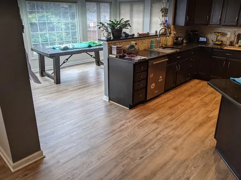 A kitchen with brown cabinets, dark countertops, and wood-look flooring. A dining table sits in a sunroom with large windows.