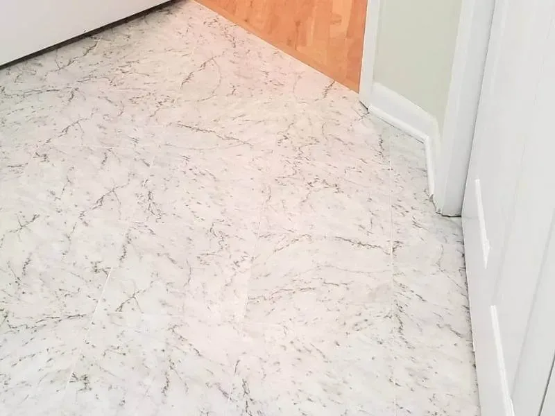 White and gray marbled patterned flooring in a room, adjacent to a white door frame and wood paneling.