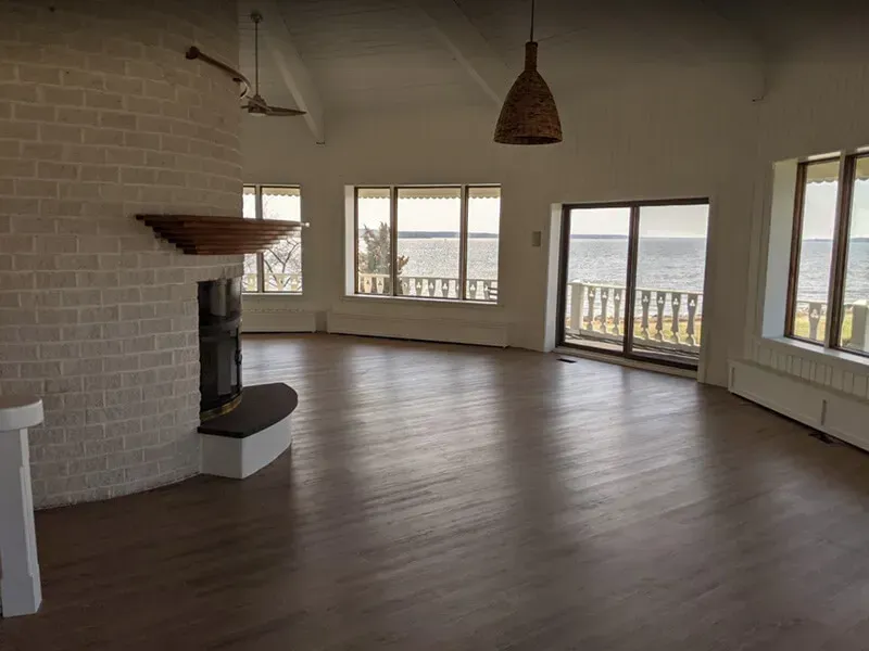 Empty living room with hardwood floors, brick fireplace, large windows overlooking the water, and a woven pendant light.