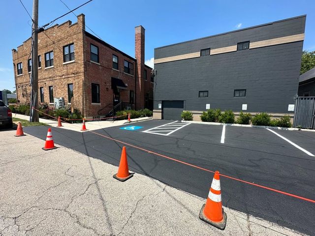 Brick building and dark gray wall beside a taped-off parking lot with orange cones on a sunny day