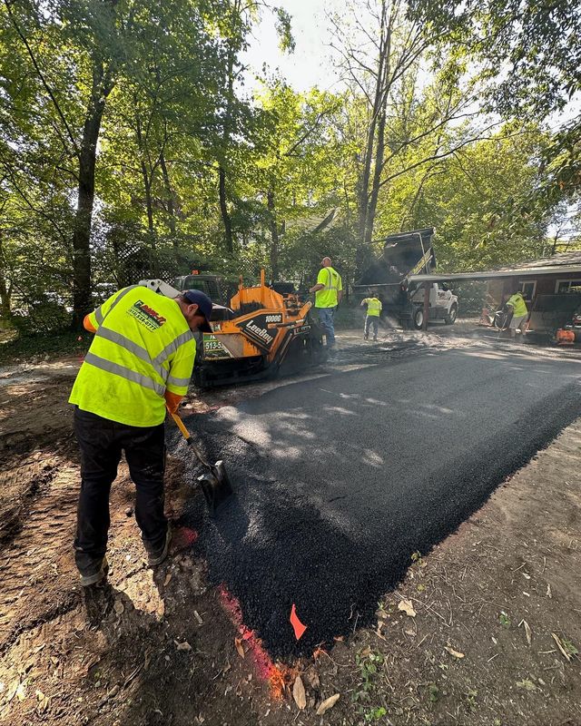 Workers paving a driveway with hot asphalt beside a small loader in a wooded yard