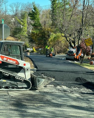 Road work with a skid-steer paving a patchy street while workers stand nearby under trees.