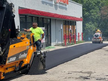Road crew paving a sidewalk beside a storefront, with an orange paving machine and fresh black asphalt.