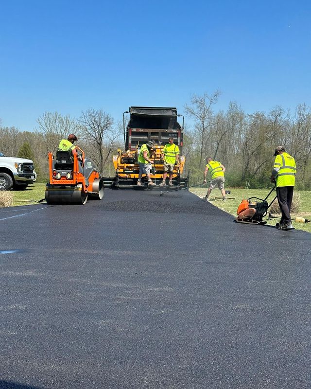 Road paving crew spreading fresh black asphalt with orange machinery under a clear blue sky.