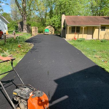 Freshly paved black driveway leading to a beige house with a red roof and trees in the yard