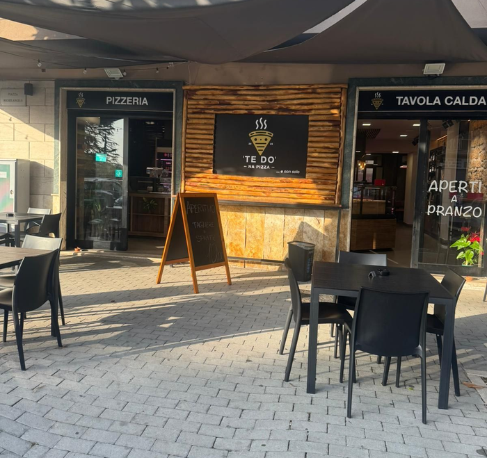 Esterno di una pizzeria e tavola calda con posti a sedere all'aperto. Tavoli e sedie neri su un patio pavimentato in mattoni.