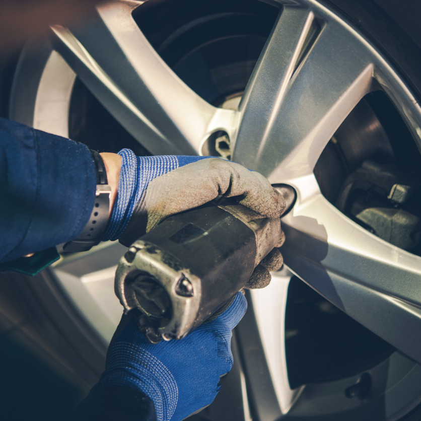 Mechanic using an impact wrench on a car tire, wearing blue gloves, in a garage.